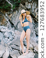 Young woman stands on a steep cliff, touching her straw hat with her hand and looking to the side 127683952