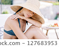 Young woman sits on a sun lounger, hugging her knees, resting her chin on her hand and looking down 127683953