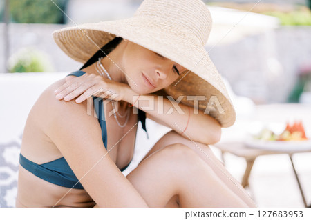 Young woman sits on a sun lounger, hugging her knees, resting her chin on her hand and looking down Young woman sits on a sun lounger, hugging her knees, resting her chin on her hand and looking down 127683953