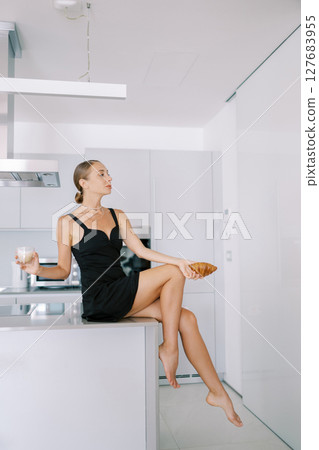 Young woman with a croissant and coffee in her hands sits on a table in the kitchen and looks away 127683955