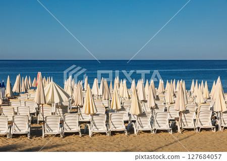 Empty beach with rows of closed sun umbrellas and white loungers facing calm blue sea under clear sky Empty beach with rows of closed sun umbrellas and white loungers facing calm blue sea under clear sky 127684057
