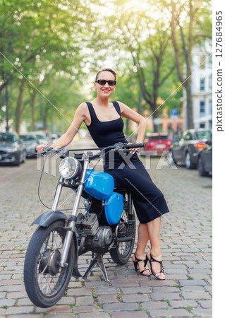 Portrait of pretty young adult stylish elegant woman wear black dress sit vintage retro blue motorcycle moped european city street Germany. Happy female person model enjoy having fun urban outside 127684965