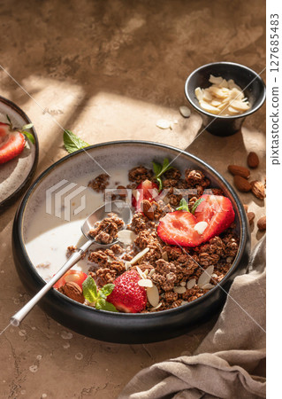 Healthy breakfast: fresh granola, muesli with milk, strawberry and almond for breakfast on brown textured background in bright morning light 127685483