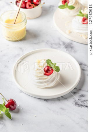 Meringues mini pavlova cake with whipped cream, fresh cherry and mint leaves on a plate, white marble background. Light summer dessert 127685618