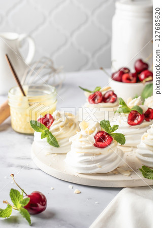 Meringues mini pavlova cakes with whipped cream decorated with fresh cherry berries and mint leaves on white marble background. Light summer dessert 127685620