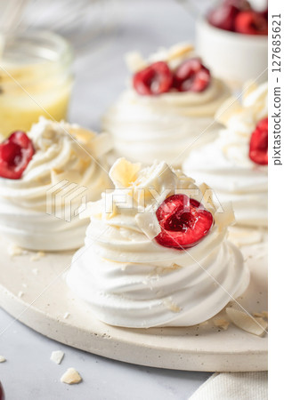 Meringues mini pavlova cakes with whipped cream decorated with fresh cherry berries on white marble background. Light summer dessert 127685621