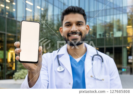 A smiling doctor holds up a phone with a blank screen, ready for your content. The medical professional stands in front of a modern building. 127685808