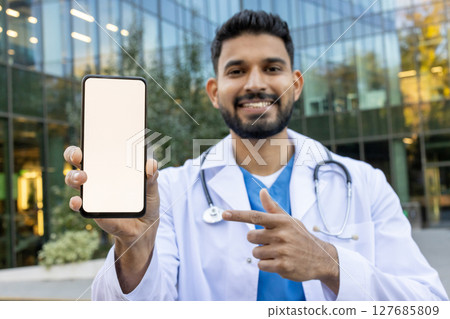 A friendly doctor holds a smartphone with a blank screen, pointing to it while smiling outside a modern building. 127685809