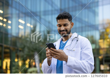 A smiling doctor wearing a white coat and stethoscope checks his phone outside a modern building. He looks happy. 127685810