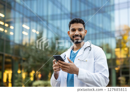 A smiling doctor looks at the camera while holding his smartphone outdoors, near a modern office building. A smiling doctor looks at the camera while holding his smartphone outdoors, near a modern office building. 127685811