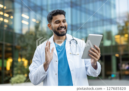 A smiling male doctor waves during a video call while holding a tablet, with a modern building in the background. A smiling male doctor waves during a video call while holding a tablet, with a modern building in the background. 127685820