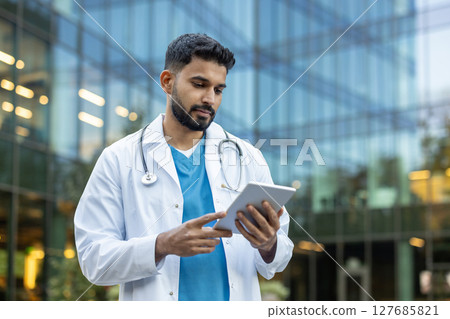 A doctor wearing a lab coat examines a tablet outside a modern medical building. He has a stethoscope around his neck. 127685821