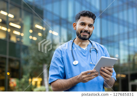 A friendly healthcare worker in blue scrubs holds a tablet while smiling, standing in front of a modern building, possibly a medical facility. 127685831