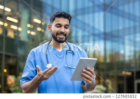 A smiling male doctor in scrubs uses a tablet outside a modern building, likely in a video call. He gestures with his hands 127685832