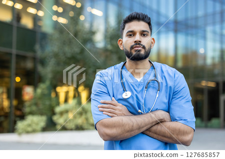 A healthcare professional stands outside, arms crossed, and is wearing scrubs and a stethoscope around his neck. The medical worker appears confident. A healthcare professional stands outside, arms crossed, and is wearing scrubs and a stethoscope around his neck. The medical worker appears confident. 127685857