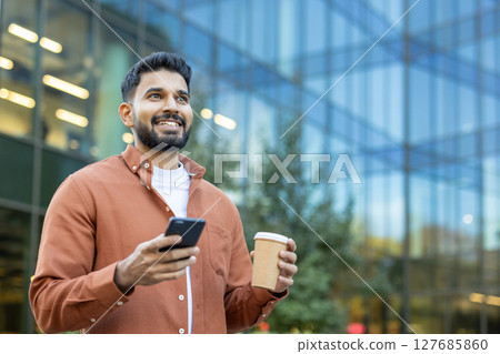 A smiling Indian man holds a coffee cup and smartphone in front of a modern glass building on a sunny day. A smiling Indian man holds a coffee cup and smartphone in front of a modern glass building on a sunny day. 127685860