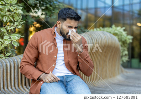 An indian man with a beard sits on a wooden bench outside, blowing his nose into a tissue during the day. The background features greenery. 127685891