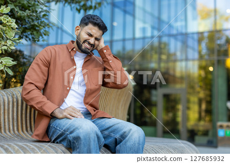 A man with a beard sitting outside, visibly distressed and clutching his ear. He appears to be experiencing otitis, an ear infection 127685932