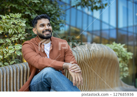 A smiling man in a casual outfit sits on a wooden bench, enjoying a sunny day outdoors with a modern building backdrop. 127685936