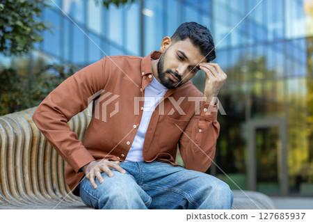 A contemplative man sits on a bench outside, deep in thought with his hand to his forehead. The modern architecture adds a sharp contrast to the scene's nature. 127685937