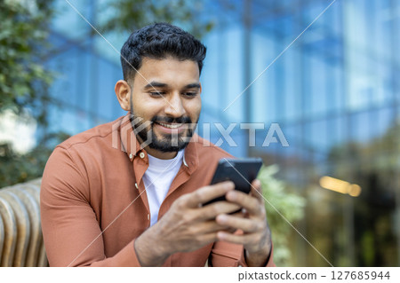 A smiling man with a beard looking at his phone while sitting outside near a modern building. 127685944