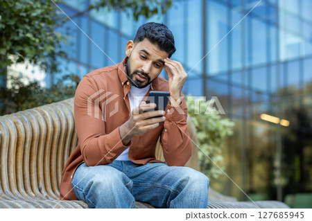 A concerned man looks at his phone while sitting outside on a bench. The scene shows a modern building in the background. 127685945