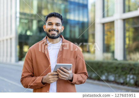 A smiling man stands outdoors, holding a tablet. He wears a casual orange shirt and a white t-shirt, with a modern building in the background. A smiling man stands outdoors, holding a tablet. He wears a casual orange shirt and a white t-shirt, with a modern building in the background. 127685951