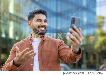 A smiling man holds up his phone, possibly video-calling or taking a selfie, outside a modern building. 127685962