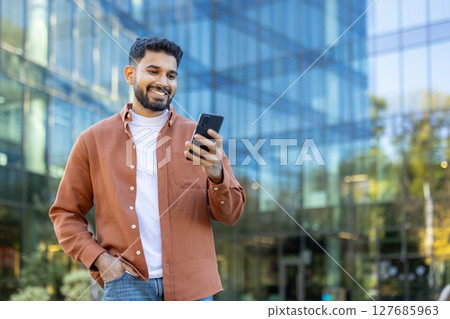 A smiling man of Indian descent looks at his phone while standing outside a modern building during the day. A smiling man of Indian descent looks at his phone while standing outside a modern building during the day. 127685963