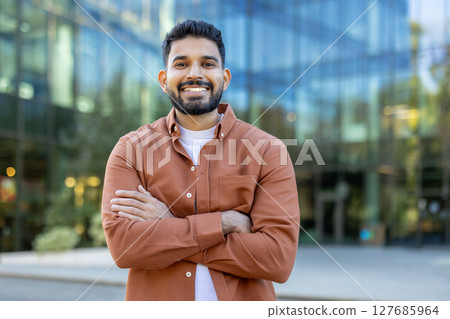 A smiling man stands confidently with arms crossed, posing in front of a modern glass building. The outdoor setting captures a professional and approachable vibe. A smiling man stands confidently with arms crossed, posing in front of a modern glass building. The outdoor setting captures a professional and approachable vibe. 127685964