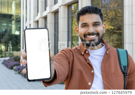 A smiling man in a brown shirt holds up a blank white smartphone screen outside a building. A smiling man in a brown shirt holds up a blank white smartphone screen outside a building. 127685979