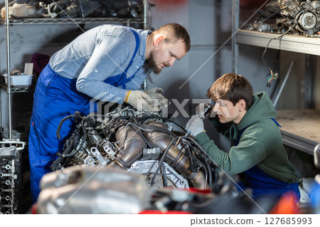 Two auto mechanics disassemble a car engine together for repair in garage Two auto mechanics disassemble a car engine together for repair in garage 127685993