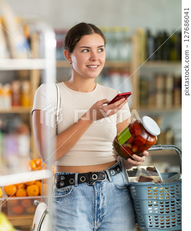 European woman customer at shop scans QR code on pickled tomatoes jar using phone camera 127686041