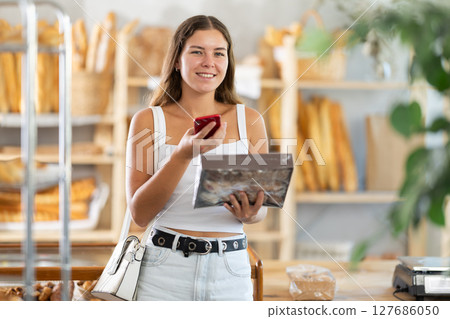 Young woman stands in bakery, scanning a cookie box with her phone Young woman stands in bakery, scanning a cookie box with her phone 127686050