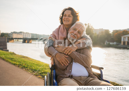 Old man in wheelchair walking with caregiver senior woman on road in park. Elderly family couple woman supporting embracing paralyzed man in chair for people with disability outdoor. Rehabilitation 127686139