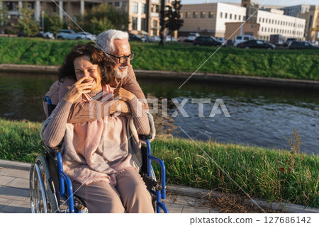 Senior woman in wheelchair walking with caregiver old man on road in park. Elderly family couple man supporting embracing woman in chair for people with disability outdoor. Rehabilitation 127686142