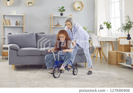 Grandmother is helping her little granddaughter ride a tricycle in the living room Grandmother is helping her little granddaughter ride a tricycle in the living room 127686906