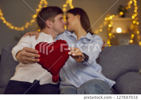 Young happy couple holding balloon heart in hands looking at each other sitting on sofa at home Young happy couple holding balloon heart in hands looking at each other sitting on sofa at home 127687018