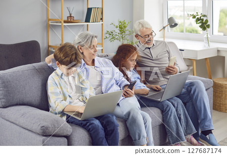 Senior grandparents resting on the couch at home with kids using laptop and smartphones together Senior grandparents resting on the couch at home with kids using laptop and smartphones together 127687174