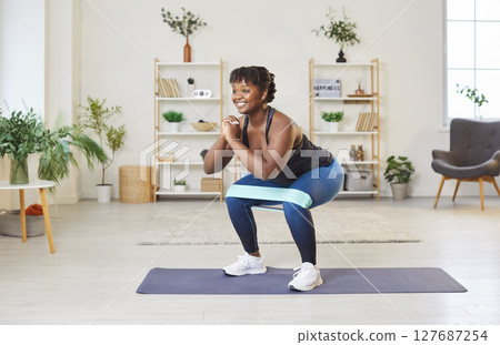 Sporty fit middle aged happy african american woman doing squat with elastic band morning exercise 127687254
