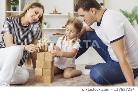 Joyful family playing together, parents and small daughter stacking wooden cubes to build tower. 127687284