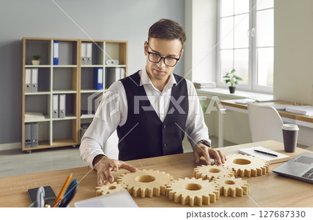 Business man connecting big wooden gears together sitting at the desk on his workplace in office. 127687330