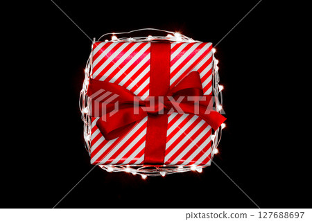 Christmas gift box with red ribbon encircled with illuminated garland and isolated on black background 127688697