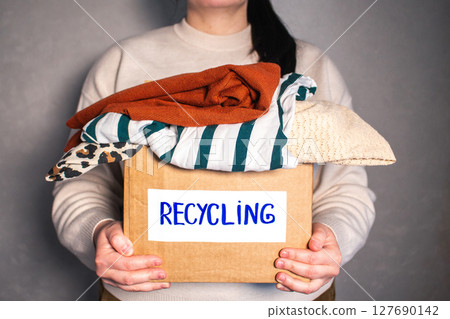 Woman holding cardboard box full of clothes labeled Recycling. Concept of sustainable fashion and responsible wardrobe management. 127690142