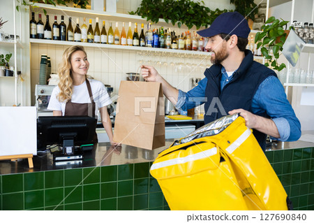 Worker at cafe counter handing order to delivery person in uniform with paper bag 127690840