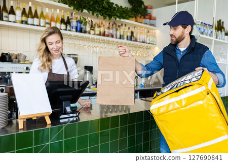 Worker at cafe counter handing order to delivery person in uniform with paper bag Worker at cafe counter handing order to delivery person in uniform with paper bag 127690841