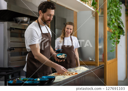 Man helps woman pizza in kitchen collaborating happily preparing meal Man helps woman pizza in kitchen collaborating happily preparing meal 127691103