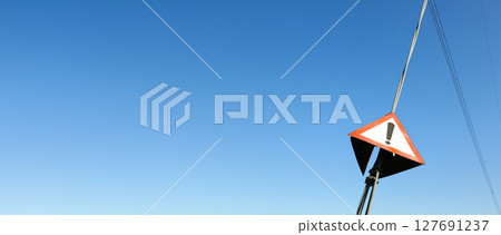 Red triangle with exclamation mark on steel cable of electric pylon, clear sky in background - wide photo, space for text left side 127691237