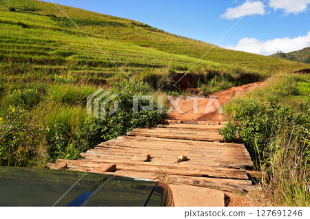 4wd car about to pass wooden bridge and red dusty road, green grass on hills landscape both sides, view from passenger window 127691246