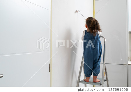 Toddler girl painting the living room wall with a roller, standing carefully on a small ladder 127691906
